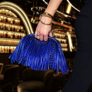 May include: A vibrant blue clutch with a unique fringed design is held by a person. The bag has a structured handle and a metallic sheen. Gold bracelets and a ring adorn the person's wrist. The background is a blurred interior.