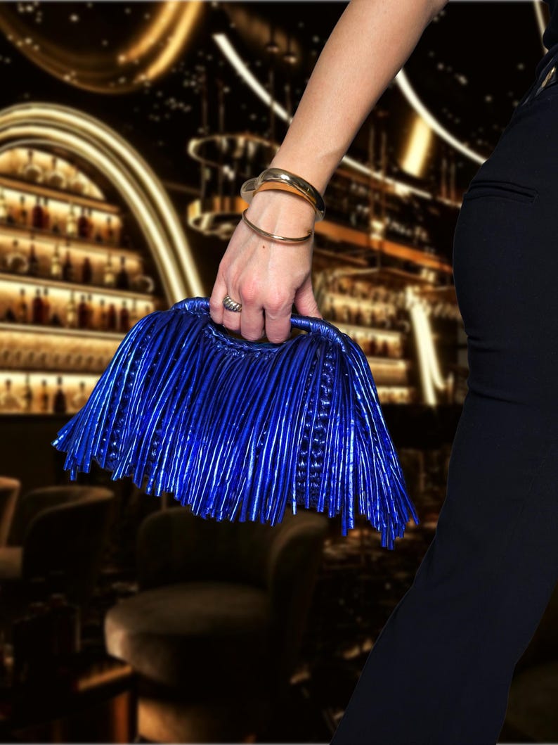 May include: A vibrant blue clutch with a unique fringed design is held by a person. The bag has a structured handle and a metallic sheen. Gold bracelets and a ring adorn the person's wrist. The background is a blurred interior.