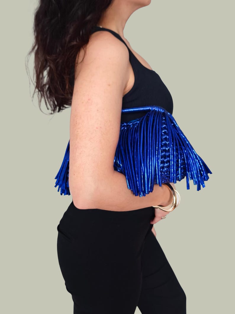 May include: A black top with a metallic blue fringe detail. The fringe is made of vertical, shiny strips. The top is paired with black pants and gold bracelets.