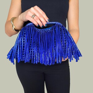 May include: A vibrant blue clutch bag with a unique fringe design. The bag is held by a person wearing a black top and pants. The clutch has a shiny, textured appearance, and the fringe adds a stylish flair.