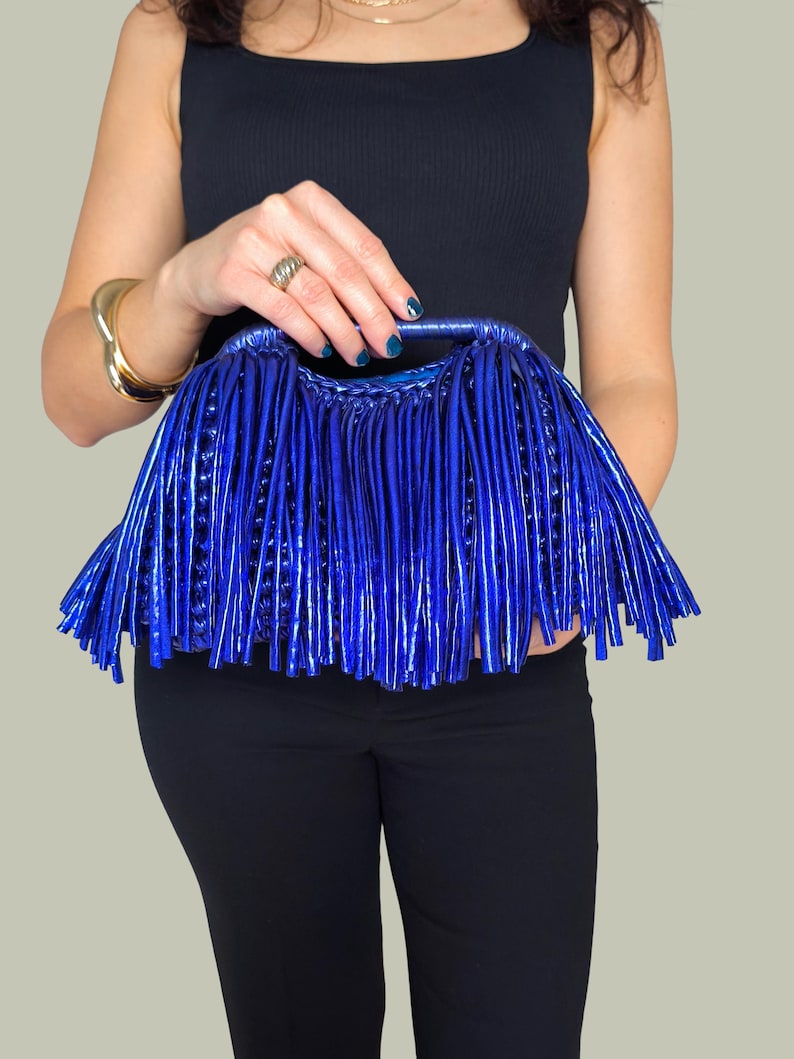 May include: A vibrant blue clutch bag with a unique fringe design. The bag is held by a person wearing a black top and pants. The clutch has a shiny, textured appearance, and the fringe adds a stylish flair.