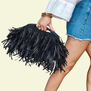 May include: A black fringed handbag with a handle. The bag is rectangular and covered in long, black fringe. The person holding the bag is wearing a white shirt and denim shorts. Gold bracelets are on the wrist.