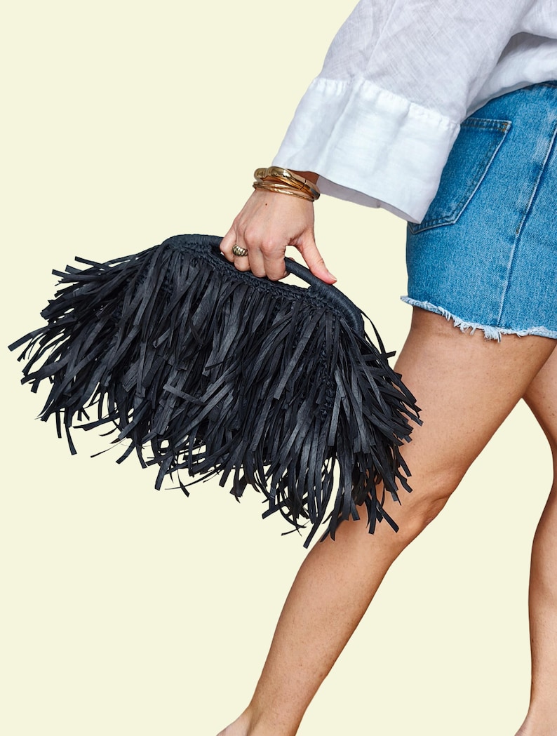 May include: A black fringed handbag with a handle. The bag is rectangular and covered in long, black fringe. The person holding the bag is wearing a white shirt and denim shorts. Gold bracelets are on the wrist.