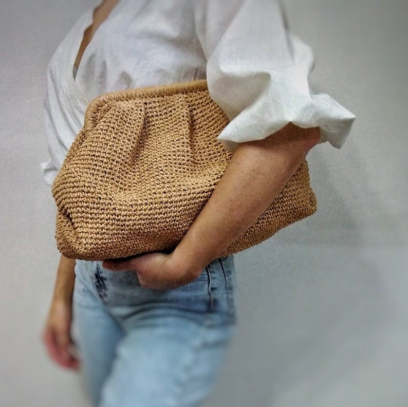 K&ouml;nnte beinhalten: Eine braune gewebte Clutch mit gerafftem Design. Die Tasche wird von einer Person gehalten, die eine wei&szlig;e Bluse und eine blaue Jeans tr&auml;gt.