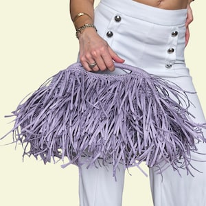 May include: A lilac fringed handbag with a woven handle. The bag is held by a person wearing white trousers and a white top. The bag's fringe is long and covers most of the bag's surface.