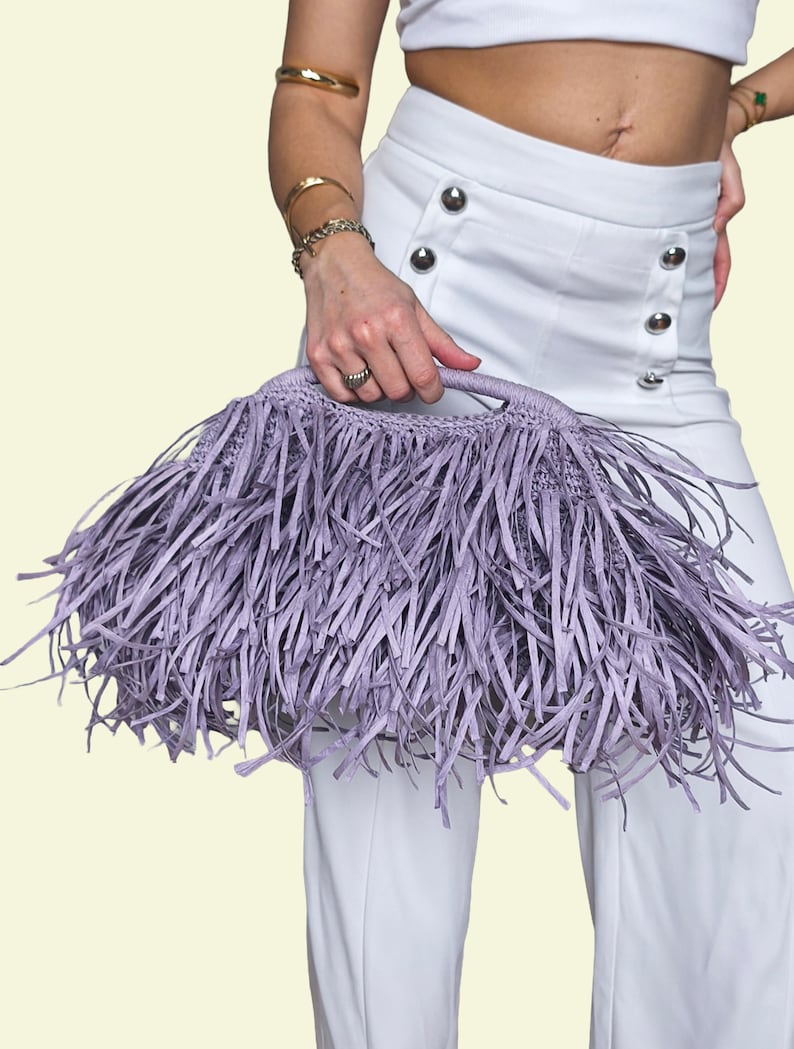 May include: A lilac fringed handbag with a woven handle. The bag is held by a person wearing white trousers and a white top. The bag's fringe is long and covers most of the bag's surface.