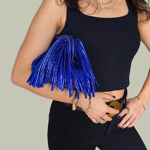 May include: A vibrant blue clutch with a fringe design. The bag is held against a black top and black pants. The clutch has a shiny, textured appearance, and is a statement accessory.