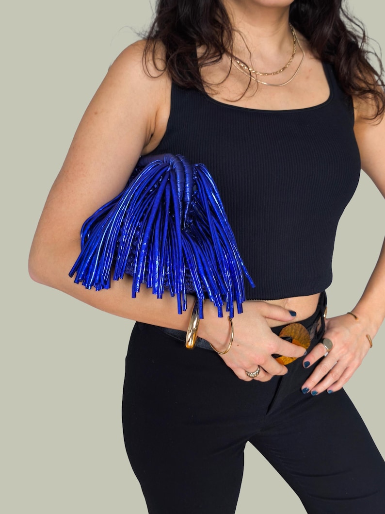 May include: A vibrant blue clutch with a fringe design. The bag is held against a black top and black pants. The clutch has a shiny, textured appearance, and is a statement accessory.