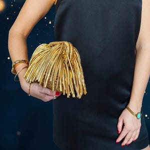 May include: A gold clutch with a fringe design is held against a black dress. The clutch has a woven base and long, metallic fringe. Gold bracelets and a watch with a turquoise stone are also visible.