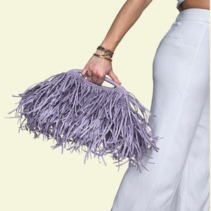 May include: A lilac fringed handbag with a handle is held by a person. The bag is made of a woven material and has long, thin fringe. The background is a light cream colour.