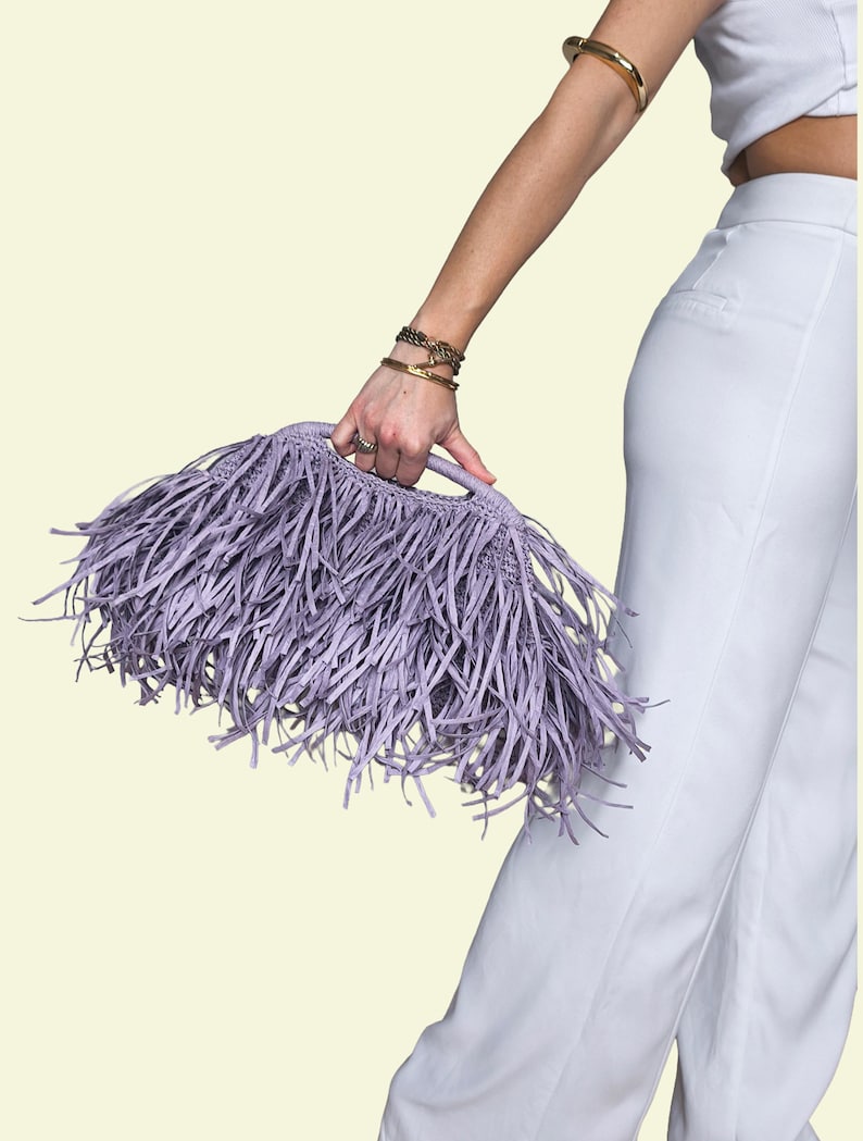 May include: A lilac fringed handbag with a handle is held by a person. The bag is made of a woven material and has long, thin fringe. The background is a light cream colour.