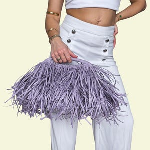 May include: A lavender fringed handbag with a woven handle. The bag is held against a background of white clothing. The bag's fringe is long and creates a textured effect. The handbag is a stylish accessory.
