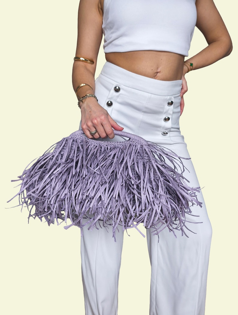 May include: A lavender fringed handbag with a woven handle. The bag is held against a background of white clothing. The bag's fringe is long and creates a textured effect. The handbag is a stylish accessory.