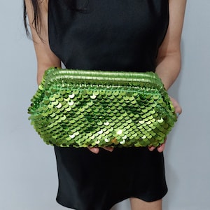 Puede incluir: Un bolso de mano verde con lentejuelas y cierre de cremallera. El bolso está hecho de pequeñas lentejuelas superpuestas que crean un efecto brillante.