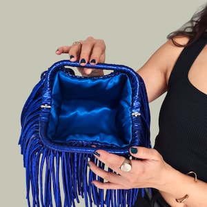 May include: A vibrant blue handbag with long fringe detailing. The bag has a structured rectangular shape with a matching blue satin interior. Silver-toned clasps secure the handles. A stylish accessory.