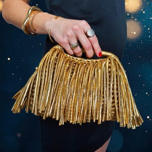 May include: A gold clutch bag with a fringe design. The bag is held by a person wearing gold bracelets and rings. The background is a dark blue with bokeh lights.