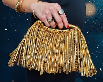 May include: A gold clutch with a fringe design. The clutch is held by a person wearing gold bracelets and rings. The background is a dark blue with bokeh lights.