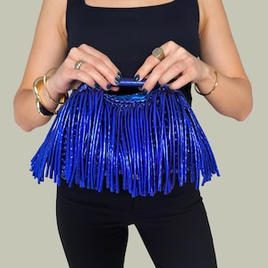 May include: A vibrant blue handbag with a fringe design. The bag has a structured handle and is held by a person wearing a black top and black pants. The bag's color is a striking, glossy blue.