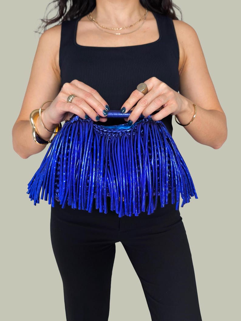 May include: A vibrant blue handbag with a fringe design. The bag has a structured handle and is held by a person wearing a black top and black pants. The bag's color is a striking, glossy blue.