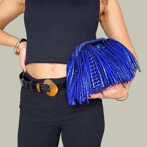 May include: A vibrant blue clutch with a unique fringe design. The bag is held against a black top and pants, with a black belt featuring a gold-colored buckle. The clutch's texture and color make it a standout accessory.