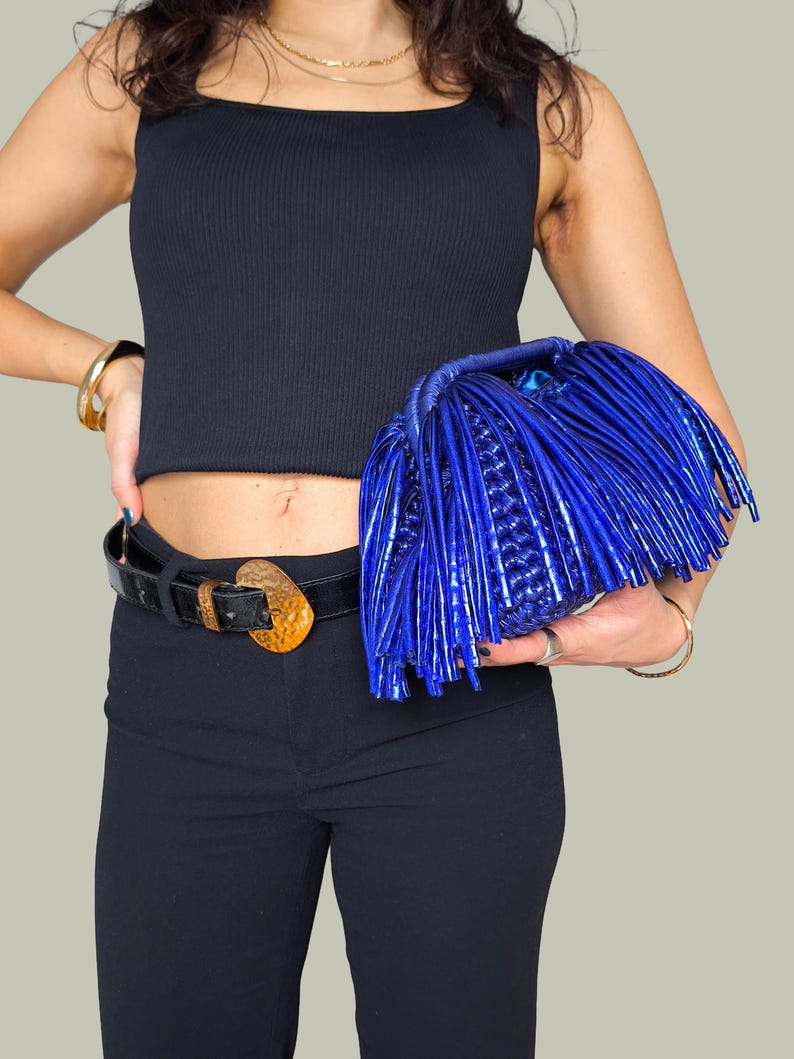 May include: A vibrant blue clutch with a unique fringe design. The bag is held against a black top and pants, with a black belt featuring a gold-colored buckle. The clutch's texture and color make it a standout accessory.