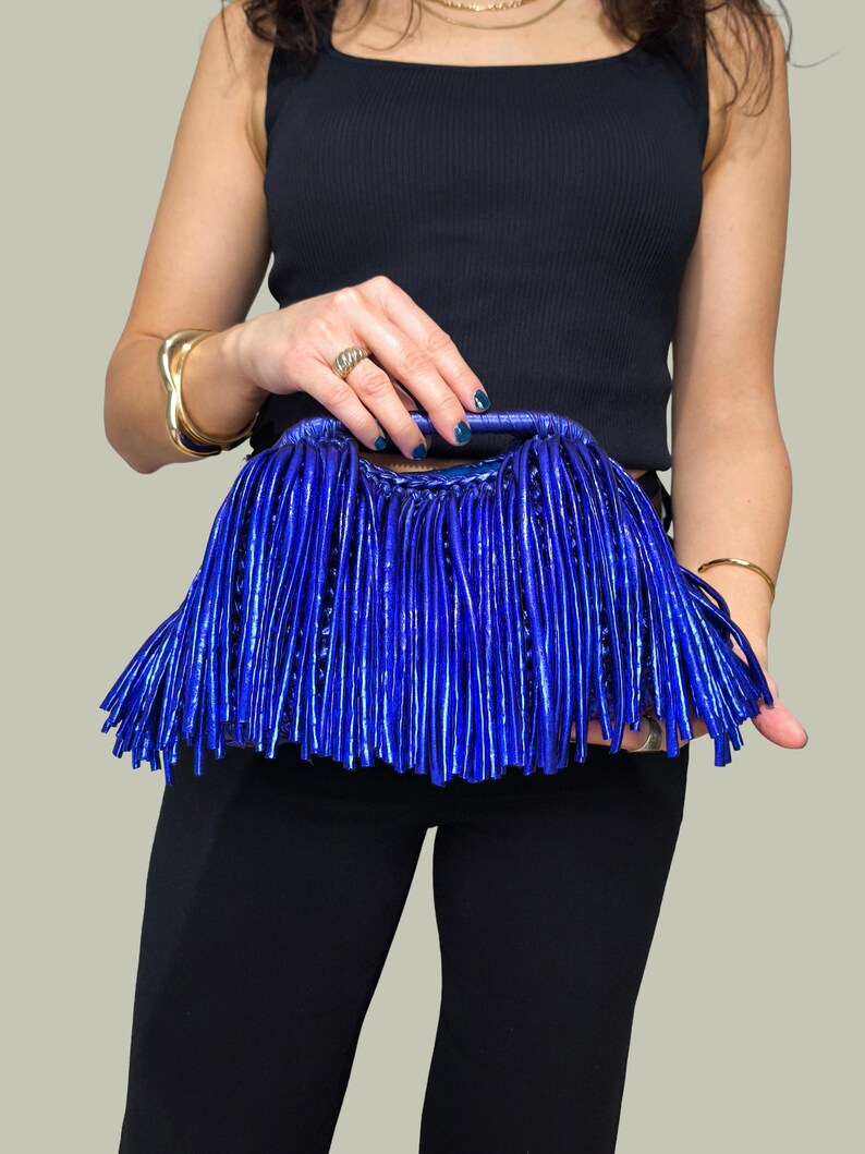 May include: A vibrant blue clutch bag with a fringe design. The bag is held by a person wearing a black tank top and black pants. Gold bracelets and a ring are also visible.