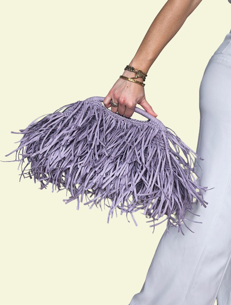 May include: A lilac handbag with long fringe detail. The bag is held by a person. The bag's handle is visible. The background is a light yellow colour.