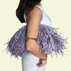 May include: A person holds a lavender fringed handbag, wearing a white top and trousers. The bag features a woven base and long, thin fringe. Gold bracelets are worn on the wrist and arm. A straw hat with a leopard print scarf is also worn.