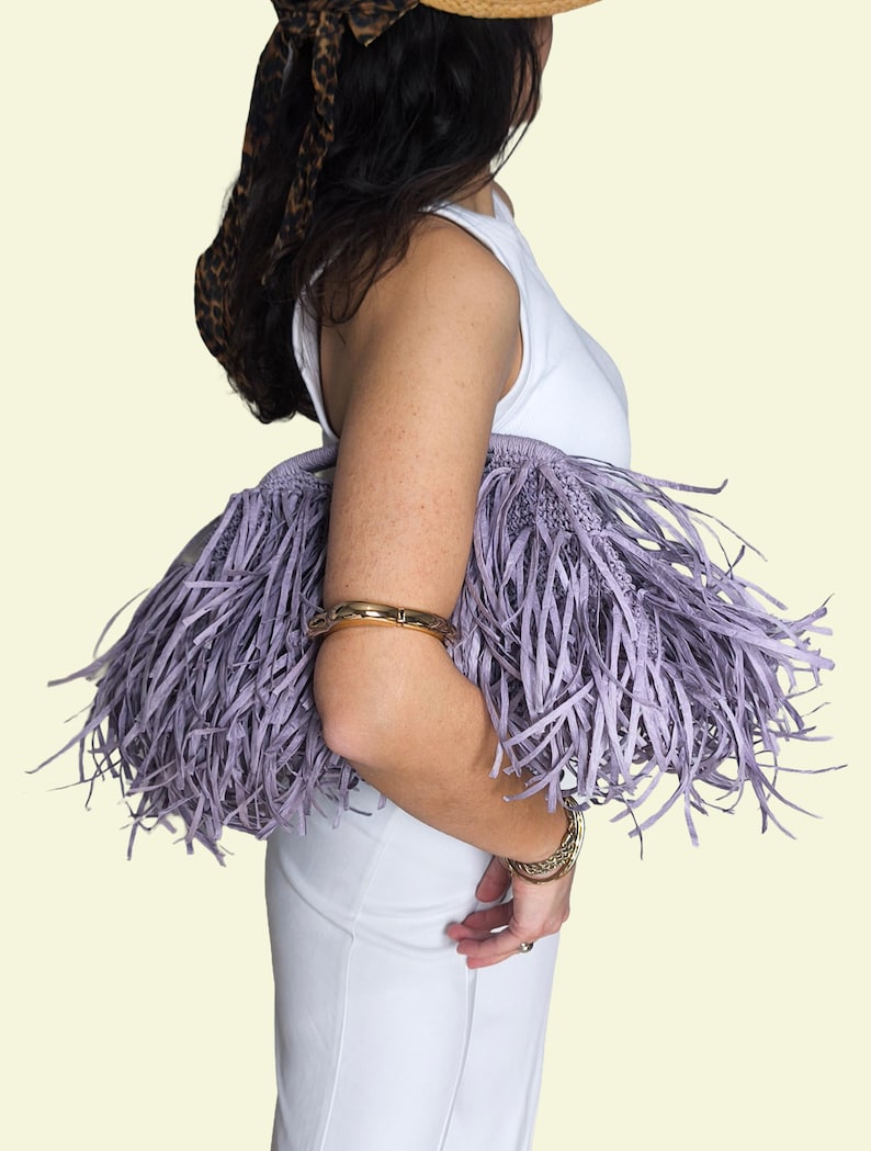May include: A person holds a lavender fringed handbag, wearing a white top and trousers. The bag features a woven base and long, thin fringe. Gold bracelets are worn on the wrist and arm. A straw hat with a leopard print scarf is also worn.