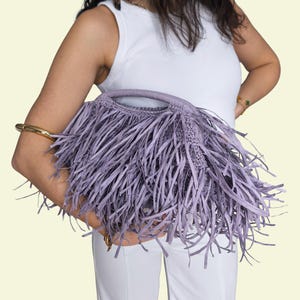 May include: A lilac-coloured handbag with a fringe design. The bag has a woven body and a handle. The fringe is long and hangs down from the bag. The bag is held by a person wearing a white vest top and white trousers.