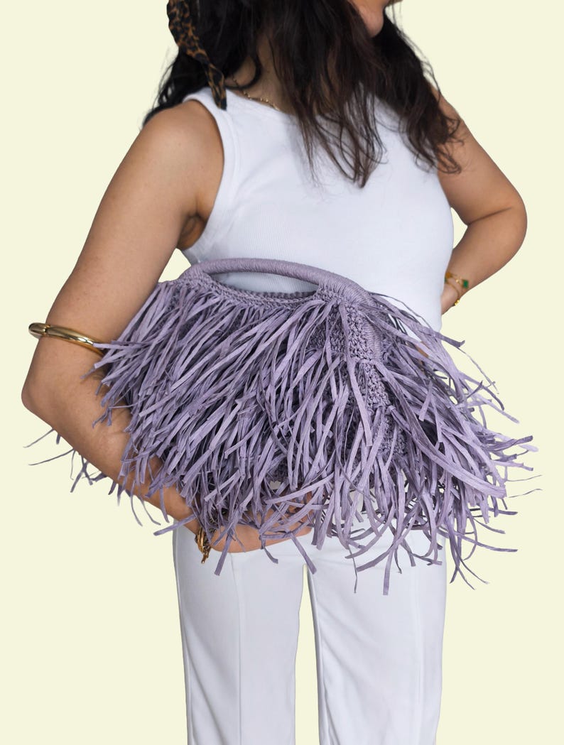 May include: A lilac-coloured handbag with a fringe design. The bag has a woven body and a handle. The fringe is long and hangs down from the bag. The bag is held by a person wearing a white vest top and white trousers.
