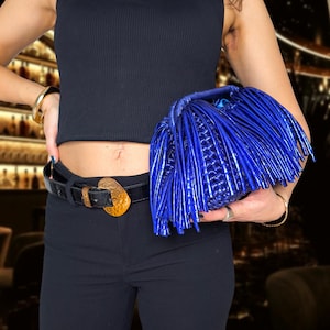 May include: A vibrant blue clutch with fringe detailing. The bag is held against a black outfit, featuring a square-neck top and black pants. A black belt with a gold-toned buckle adds a contrasting accent. Gold jewelry completes the look.