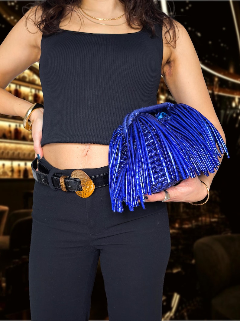 May include: A vibrant blue clutch with fringe detailing. The bag is held against a black outfit, featuring a square-neck top and black pants. A black belt with a gold-toned buckle adds a contrasting accent. Gold jewelry completes the look.