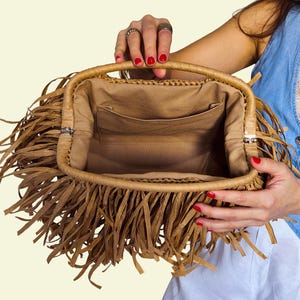 May include: A tan handbag with a rectangular shape and a woven handle. The bag has a fringe detail around the edges and a tan fabric interior with a pocket. The bag is held open, showing the inside.