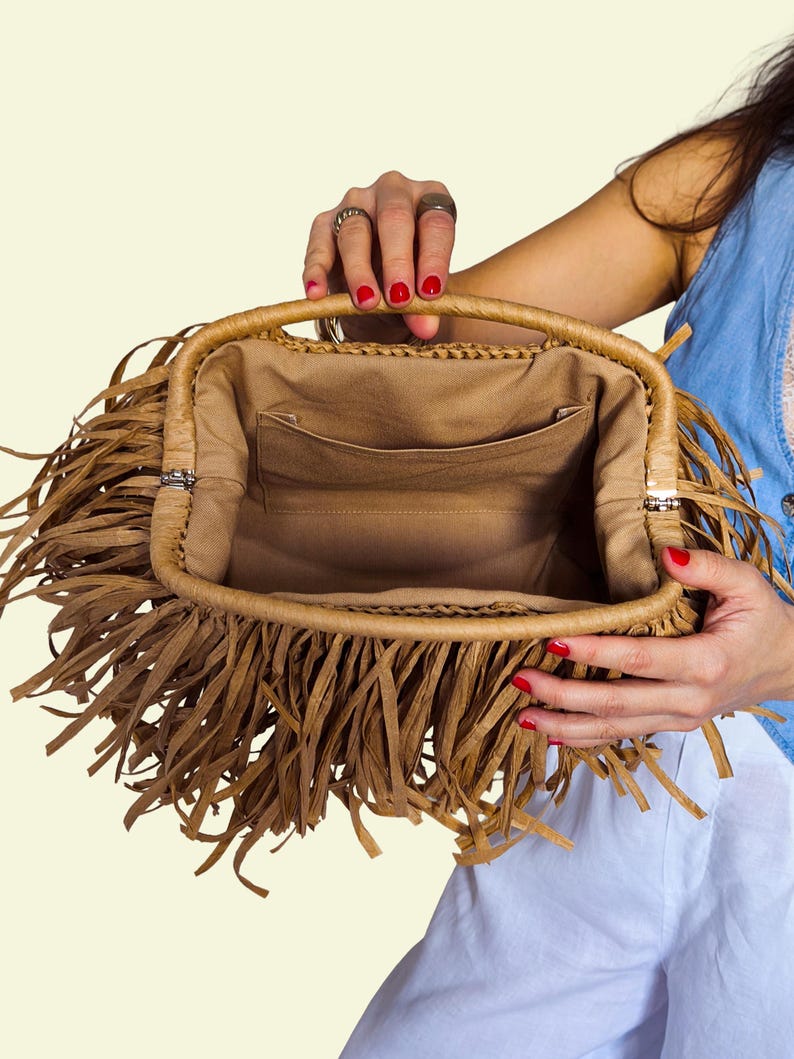 May include: A tan handbag with a rectangular shape and a woven handle. The bag has a fringe detail around the edges and a tan fabric interior with a pocket. The bag is held open, showing the inside.