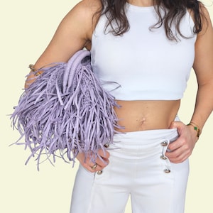 May include: A lilac handbag with a fringed design. The bag has a woven structure and two handles. The model is wearing a white vest top and white trousers with silver buttons. The background is a light yellow colour.