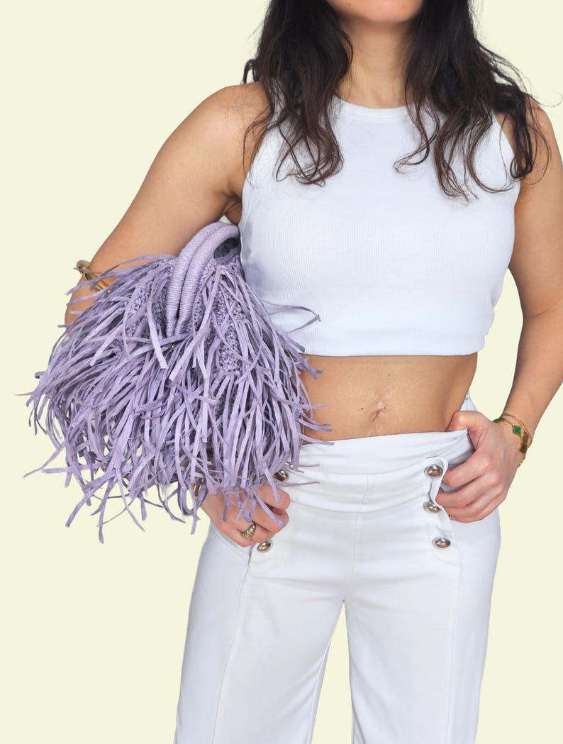 May include: A lilac handbag with a fringed design. The bag has a woven structure and two handles. The model is wearing a white vest top and white trousers with silver buttons. The background is a light yellow colour.