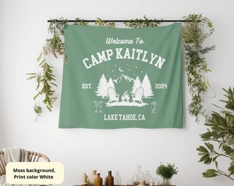Op de afbeelding: Een groene wandkleed met witte tekst die "Welkom bij Camp Kaitlyn Est. 2024 Lake Tahoe, CA" luidt. De wandkleed toont een kampvuurscène met twee stoelen en een bergketen op de achtergrond.