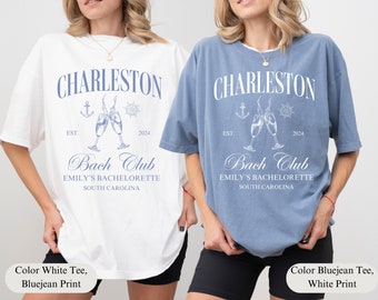 May include: Two white t-shirts with blue text and graphics. The text reads "Charleston Bach Club Emily's Bachelorette South Carolina" with the years "EST. 2024". One shirt is white with blue text and graphics, the other is blue with white text and graphics.