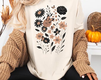 May include: Ivory-colored t-shirt featuring a floral and butterfly design in black and peach tones. The graphic includes various flower shapes and butterflies. The word "IVORY" is printed in a black, retro font in the upper right corner.