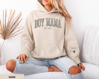 Op de afbeelding: Een zandkleurige hoodie met de tekst "BOY MAMA EST. 2026" in een patroonlettertype over de borst. De mouw heeft de naam "Chris" en een hart. De hoodie heeft een voorzak en een capuchon.