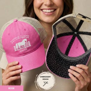 Op de afbeelding: Roze truckerpet met een wit geborduurd ontwerp met een paard en de tekst "Dutton Ranch". De pet heeft een mesh achterkant en een zwart-wit stippenpatroon aan de onderkant van de klep. De tekst "ESTD. 2016" is ook zichtbaar.