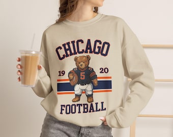 Puede incluir: Una sudadera beige de cuello redondo con un gráfico de un oso de peluche vestido con un uniforme de fútbol americano azul oscuro y naranja con el número 5, sosteniendo un balón. El texto CHICAGO y FOOTBALL rodea al oso, junto a los números 19 y 20.