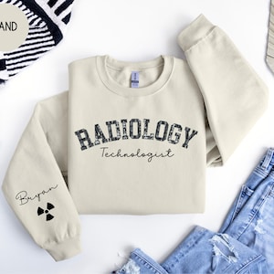 Może przedstawiać: Szarobrązowa bluza z nadrukiem "RADIOLOGY Technologist" na przodzie. Lewy rękaw ma czarny symbol promieniowania i napis "Bryan" nadrukowany na nim.
