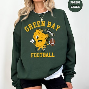 May include: Forest green sweatshirt with the words "GREEN BAY FOOTBALL" in yellow. A cartoon cheese character is holding a football. The sweatshirt has a white undershirt peeking out from the neckline.