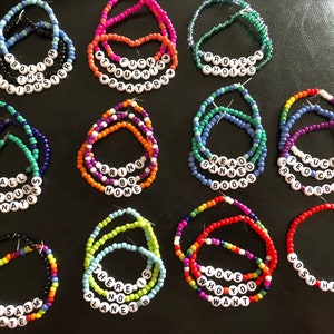 May include: A collection of 12 colorful beaded bracelets with various messages, including "Abolish the Filibuster", "Read Banned Books", "Love Who You Want", and "Disarm Hate".