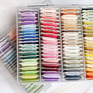 May include: A clear plastic storage box filled with rows of colourful embroidery floss spools. Each spool is labelled with a number and letter code, such as A337, A363, and A822.