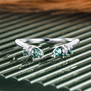 May include: A silver ring with a split shank design. The ring features two clusters of green gemstones, each resembling leaves. The gemstones are marquise and round cut. The ring is set on a green and brown surface.