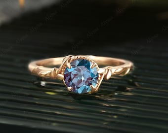 Puede incluir: Un anillo de compromiso de oro rosa con una piedra preciosa de alexandrita azul y verde engastada en una banda delicada y retorcida.