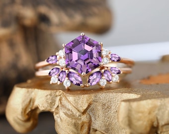 Puede incluir: Un anillo de compromiso de oro rosa con una gran piedra central de amatista hexagonal rodeada de pequeños acentos de amatista y diamantes. El anillo está colocado sobre un soporte dorado.
